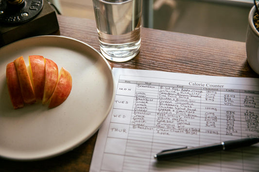 Overhead view of a handwritten meal log on a desk beside a sliced apple and a glass of water, symbolizing mindful eating and habit awareness.
