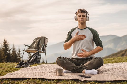 Man sitting on a blanket outdoors wearing over-ear headphones, eyes closed, one hand on chest and one hand on belly practicing mindful breathing meditation