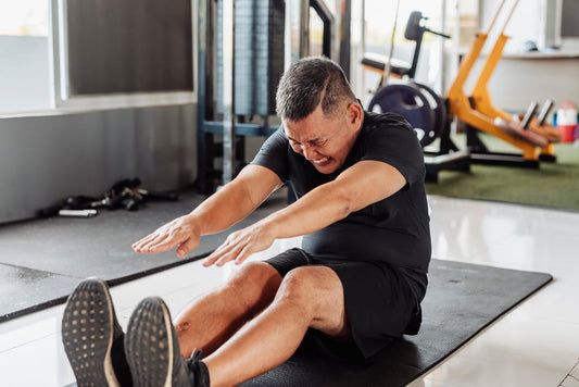 man on gym mat reaching for his feet with a pained expression, incorrectly flexing his lower back during a hamstring stretch