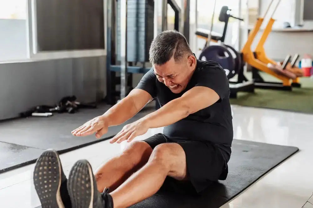 man on gym mat reaching for his feet with a pained expression, incorrectly flexing his lower back during a hamstring stretch
