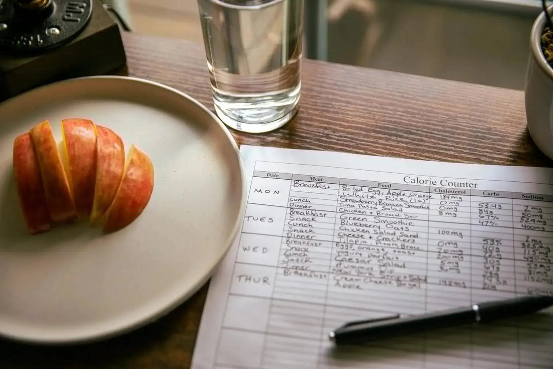 Overhead view of a handwritten meal log on a desk beside a sliced apple and a glass of water, symbolizing mindful eating and habit awareness.