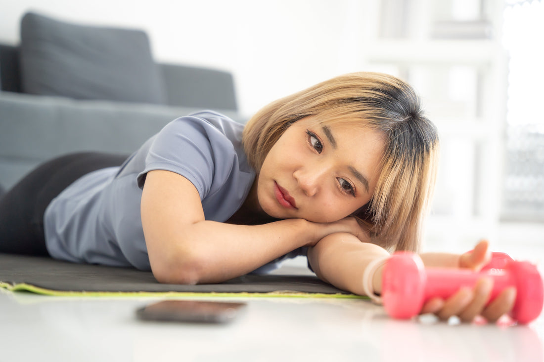 Woman lying on the floor holding a dumbbell and staring at it, looking tired and unmotivated before a workout.