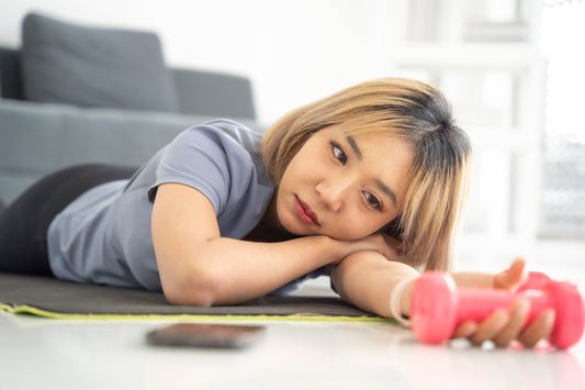 Woman lying on the floor holding a dumbbell and staring at it, looking tired and unmotivated before a workout.