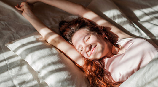 Smiling woman stretching in bed as morning sunlight streams through window blinds, illustrating how morning light supports circadian rhythm and daytime energy.