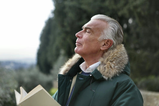 Older gentleman standing outdoors with closed eyes, holding a book while taking a deep breath of fresh air in a natural setting.