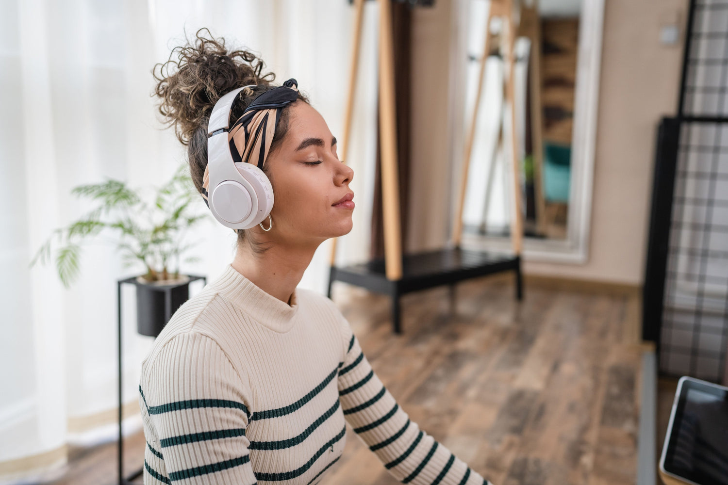  A young woman sitting cross-legged indoors, meditating with headphones on, eyes closed, and a peaceful expression, surrounded by a calm and minimalist setting.